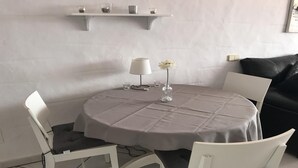 A round table with a grey tablecloth, two white chairs, a vase with a single flower, a floor lamp, and a shelf above holding a few items.
