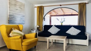Una habitación con un sillón amarillo, un sofá azul oscuro con cojines estampados, una pequeña mesa de madera y una ventana con cortinas.