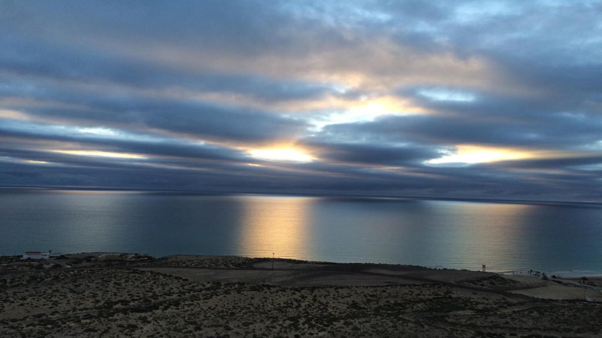 A coastal landscape with a calm sea, overcast sky, and a barren terrain in the foreground.