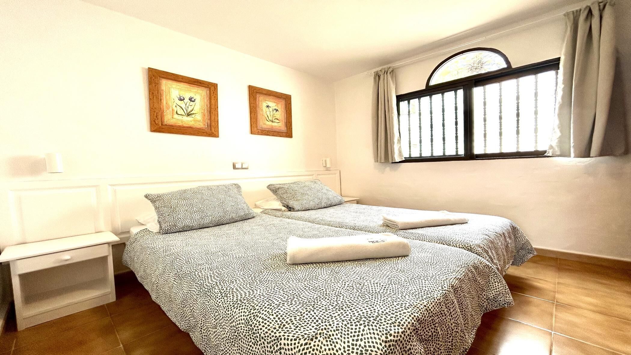 A hotel room with two beds, a nightstand, a window with curtains, and two framed pictures on the wall.