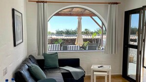 Une pièce avec un canapé en cuir noir, une petite table blanche et une fenêtre donnant sur un balcon et des plantes d’extérieur.