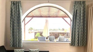Une pièce dotée d’une grande fenêtre en arc de cercle, d  ’une petite table avec deux chaises, et offrant une vue sur un jardin et des bâtiments à l’extérieur.