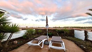 Une terrasse dotée de deux chaises longues et d’un parasol, donnant sur un espace aménagé avec un mur en pierre et offrant une vue sur un champ situé au loin.