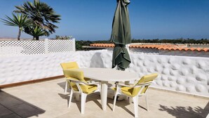 Un patio con una mesa blanca y sillas amarillas, una sombrilla verde y vistas al mar.
