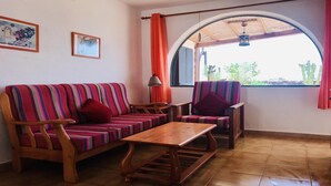 Ein Wohnzimmer mit einem gestreiften Sofa, einem Holztisch und einem Sessel. An der Wand hängen zwei Gemälde, ein Fenster bietet Blick auf Grünflächen, und ein Deckenventilator ist angebracht.