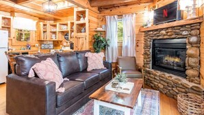 A cozy cabin interior with a fireplace, wooden walls, a leather sofa, and a kitchen area.