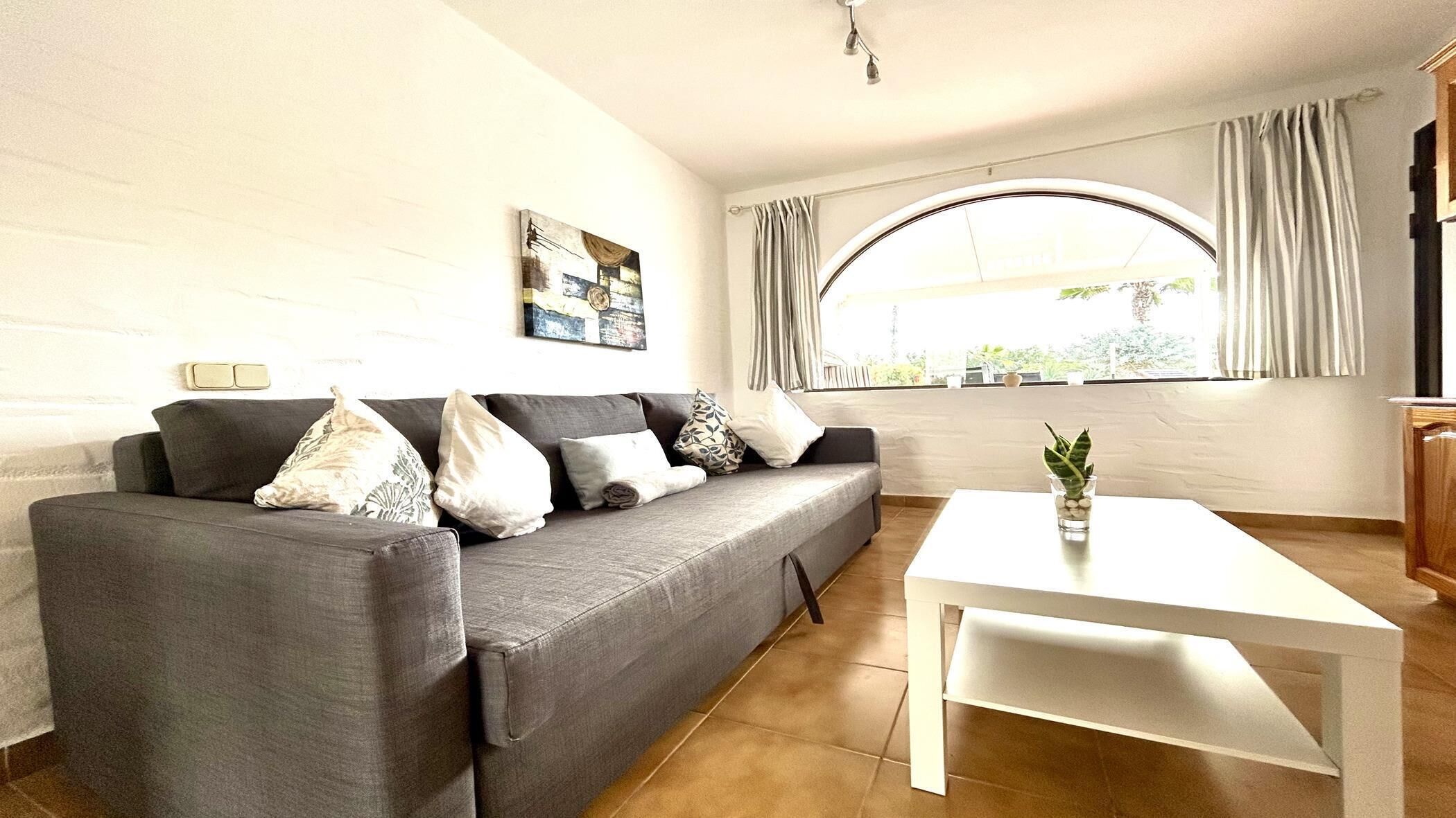 Uma sala de estar com sofá cinzento, almofadas brancas, mesa de centro branca e janela com cortinas.
