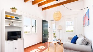 Un salon avec un téléviseur posé sur un meuble blanc, un canapé avec des coussins, une petite table avec une lampe et un luminaire suspendu.