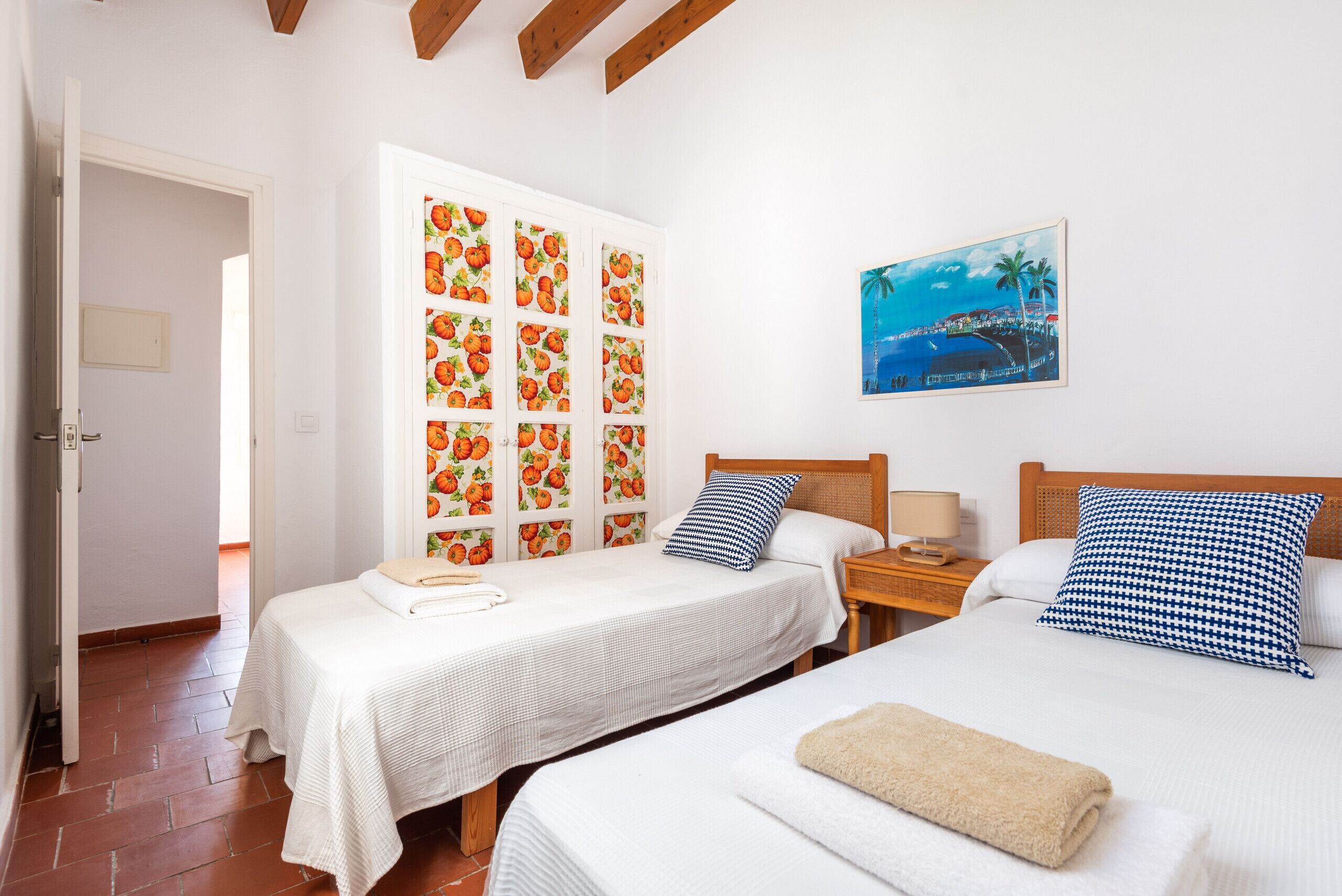 A hotel room with two beds, a painting of a coastal scene, and a wall with fruit-patterned panels.