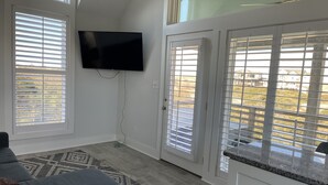 A modern living room with a grey sofa, a flat-screen TV mounted on the wall, and a large sliding glass door leading to a balcony. - Romantic Little Getaway in 4X4 beach (Corolla)