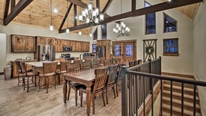 A spacious kitchen with wooden cabinets, a large island, and modern appliances. There is a dining area with wooden tables and chairs, and a staircase leading to an upper level. - Lake View | Game Room | King Beds | Hot Tub | Pool (Branson)