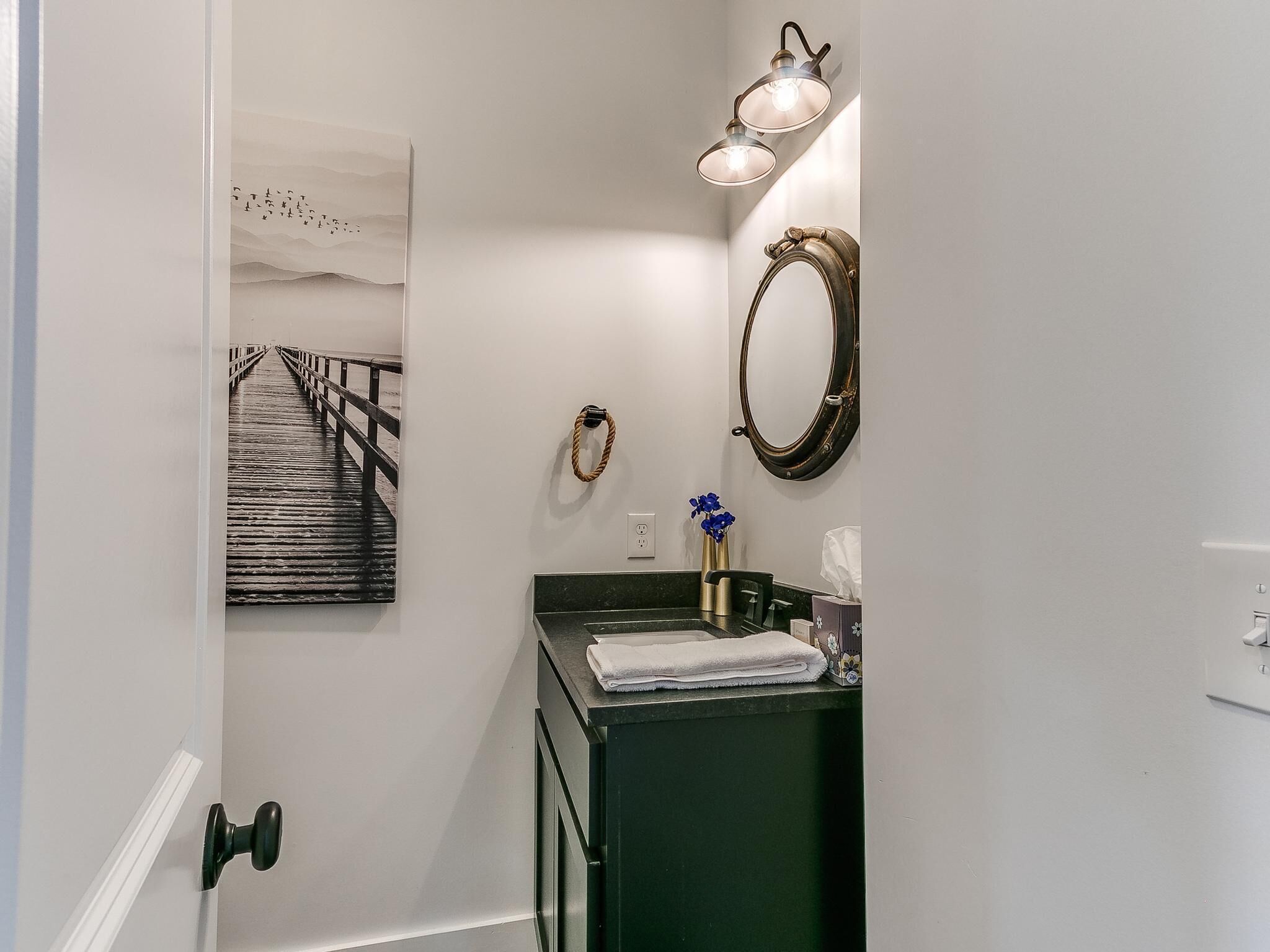 A bathroom with a wooden floor, a black vanity, a mirror, and a painting of a bridge.