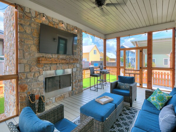 A covered outdoor patio with stone walls, a fireplace, wicker furniture, and a TV mounted on the wall.