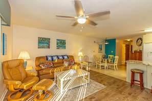 A living room with a brown leather sofa, a coffee table, a ceiling fan, and a dining area with chairs and a table.