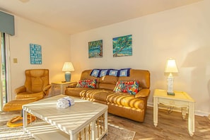 A living room with a brown leather sofa, two armchairs, a coffee table, and two side tables with lamps. There are two paintings on the wall and a sign with text.