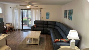 A living room with a sectional sofa, a coffee table, a lamp, a rug, a ceiling fan, and a sliding glass door leading to an outdoor area. - Golf Colony at Deerfield 33-I (Myrtle Beach)
