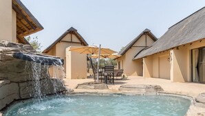 Zona de piscina con cascada, mobiliario exterior y edificios con techo de paja.