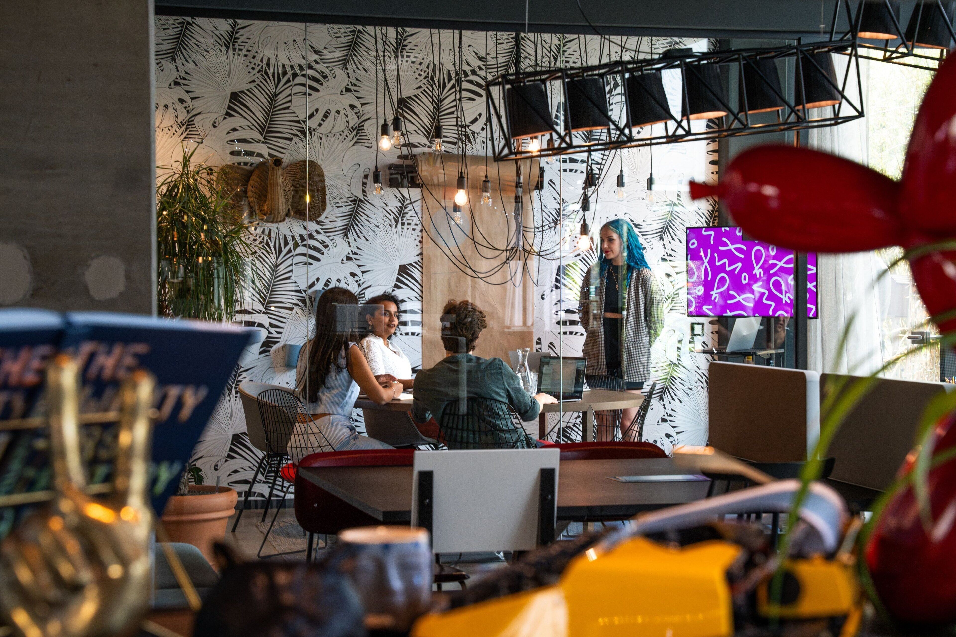Un espacio de oficina moderno con un área de reuniones colaborativa, un ventanal grande y una pared decorativa con motivos de hojas.