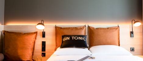 Habitación de hotel con cama, una guitarra, almohada con el texto “GIN TONIC” y dos lámparas de pared.