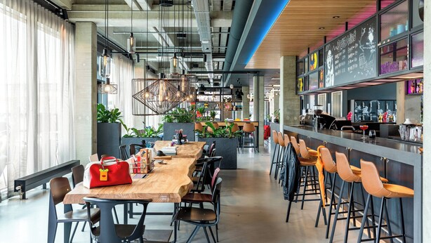 A modern bar area with a long wooden counter, bar stools, and a chalkboard menu. - Moxy Utrecht (Utrecht)