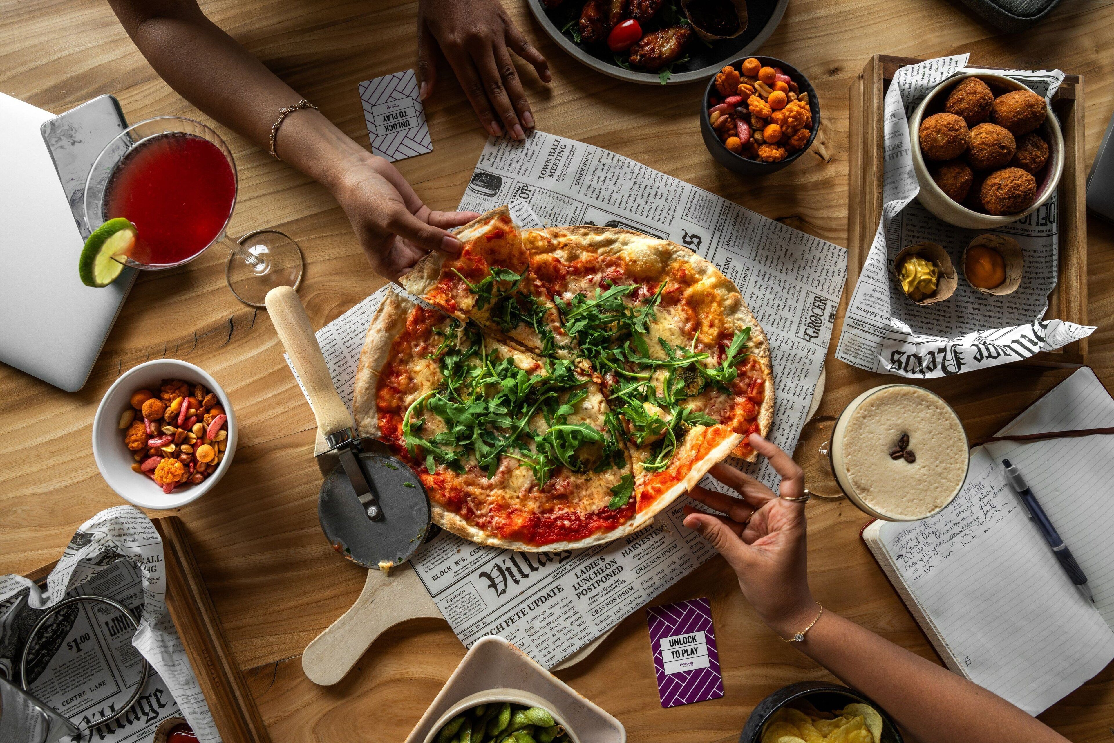 Pizza op een houten tafel, onder ligt kranten, omringd door verschillende gerechten en een laptop.