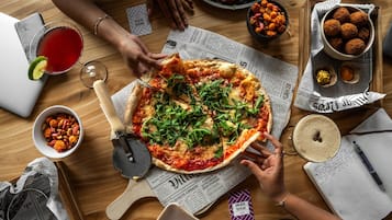 Pizza op een houten tafel, onder ligt kranten, omringd door verschillende gerechten en een laptop.