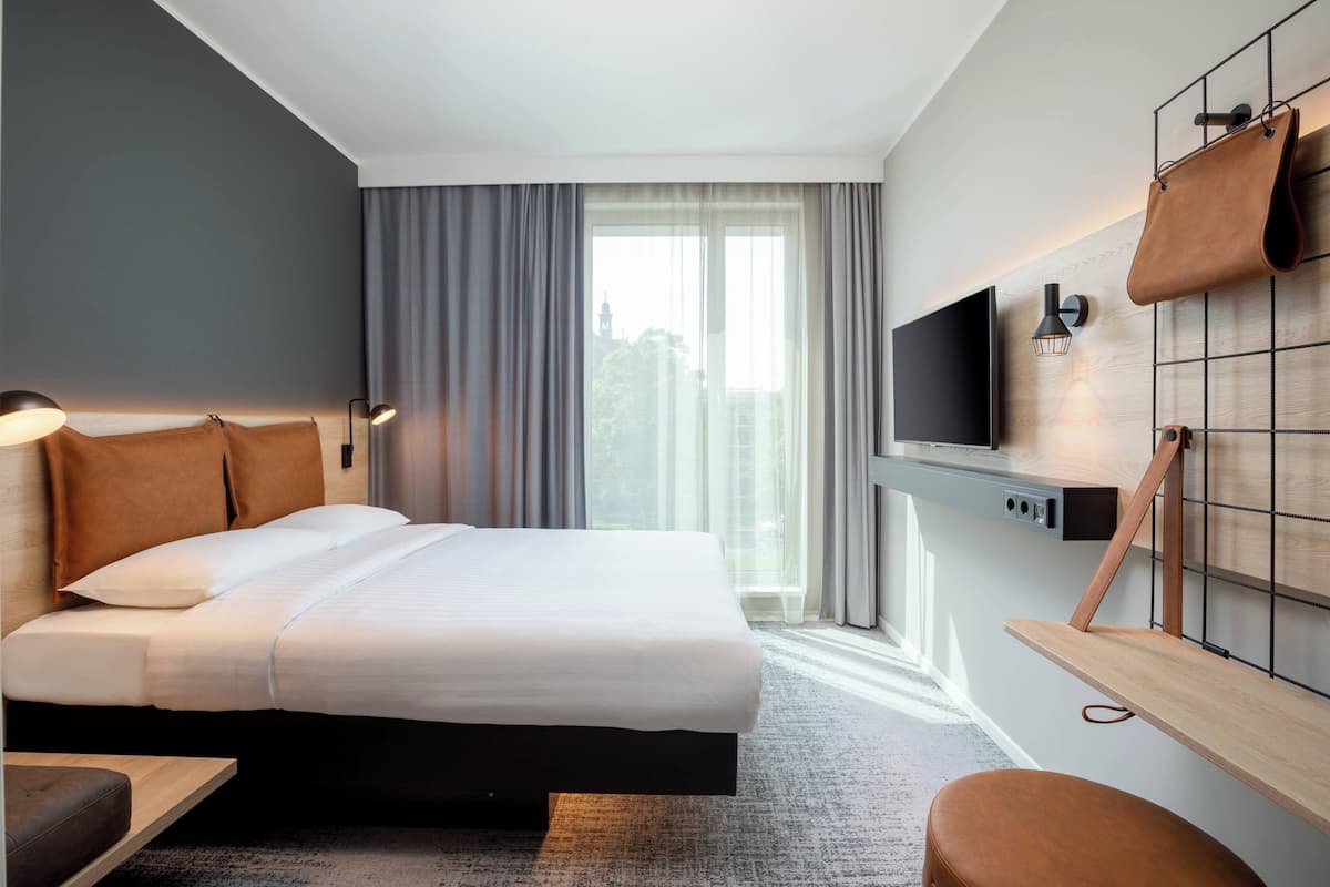 Habitación de hotel moderna con una cama grande, un televisor montado en la pared, un escritorio y vistas a la ciudad a través del ventanal.