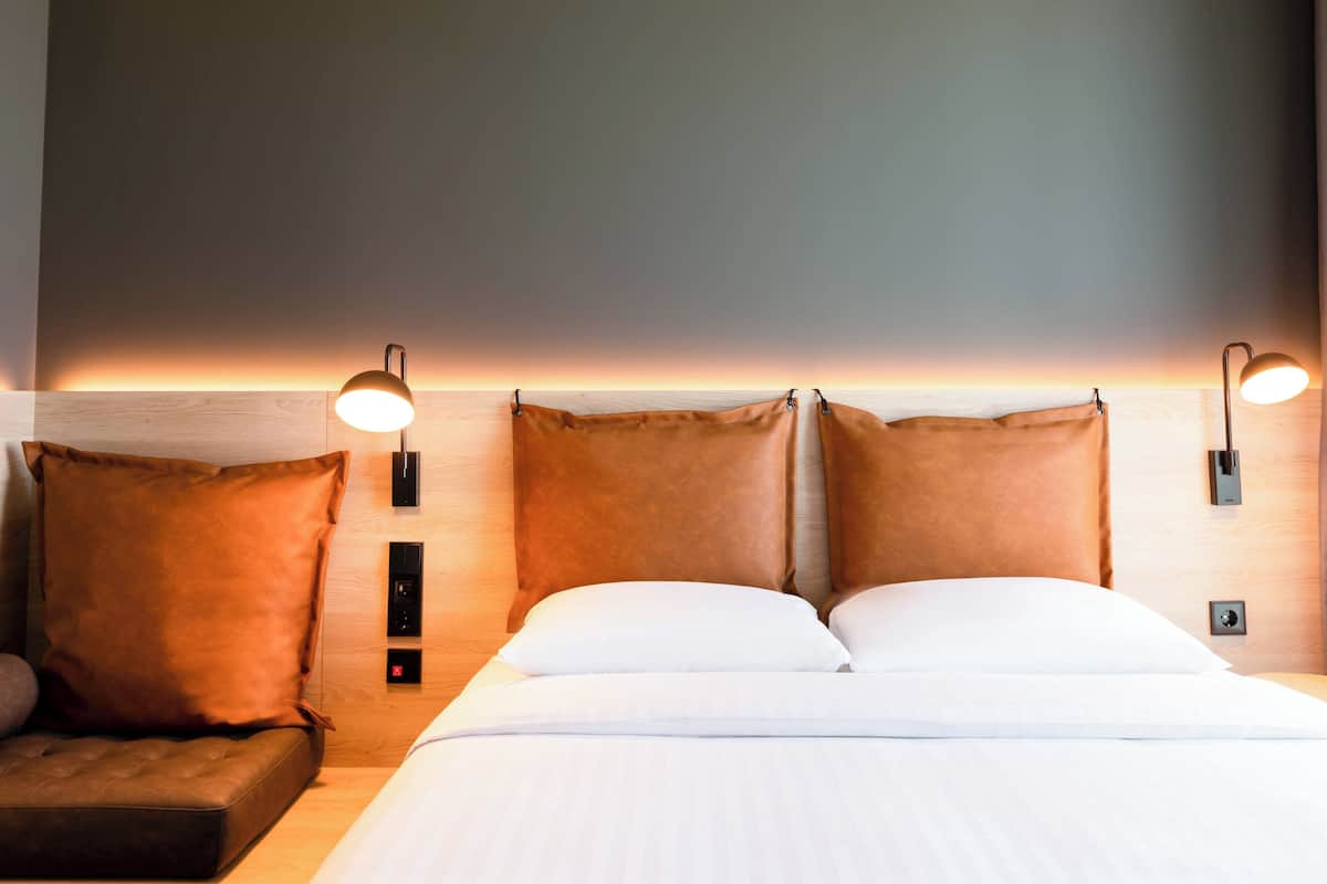 Una cama bien hecha con sábanas blancas y dos almohadas marrones, con dos lámparas fijadas a la pared a cada lado.