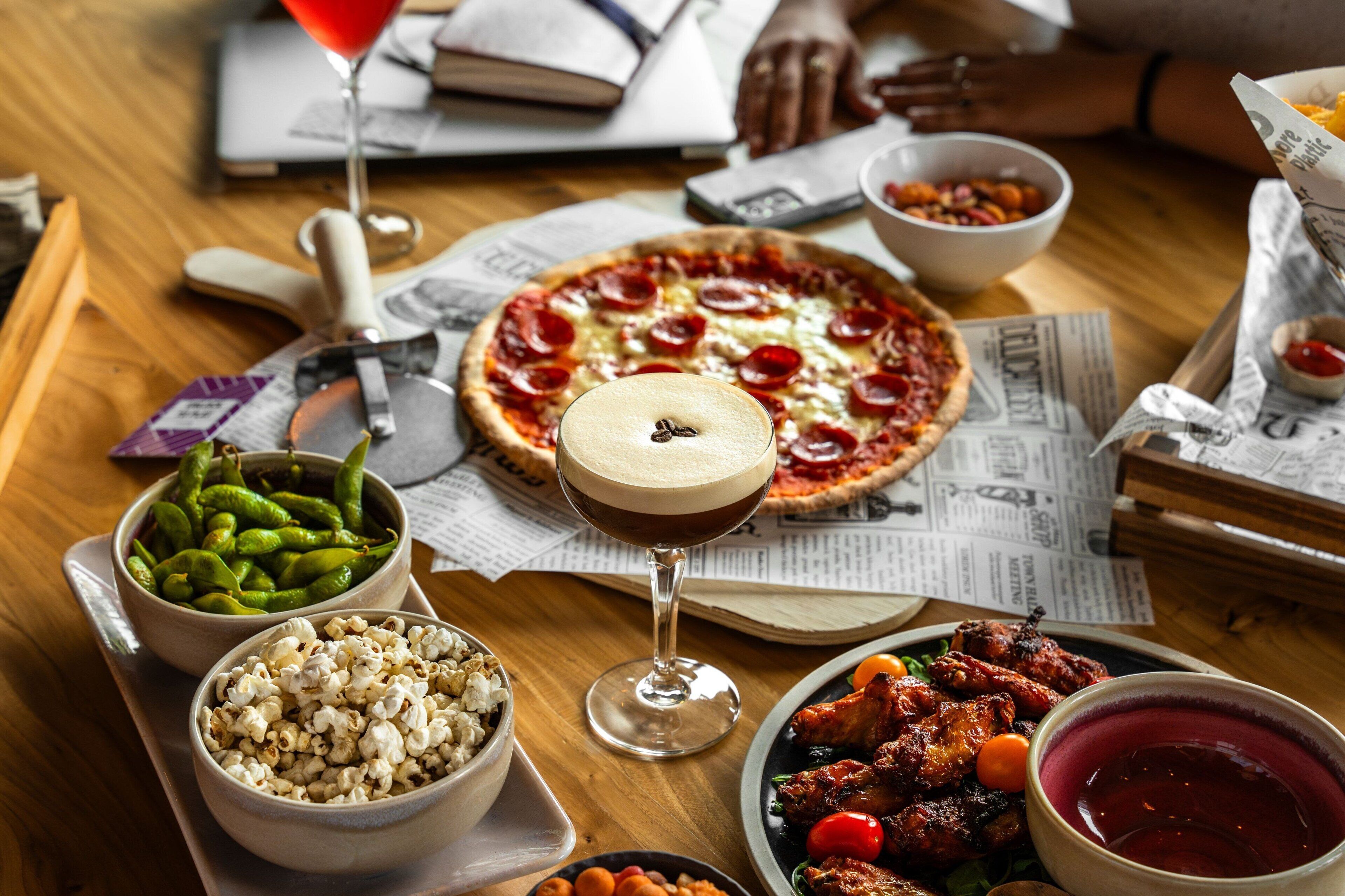 Uma mesa com pizza, pipoca, uma taça de martini e diversos acompanhamentos.