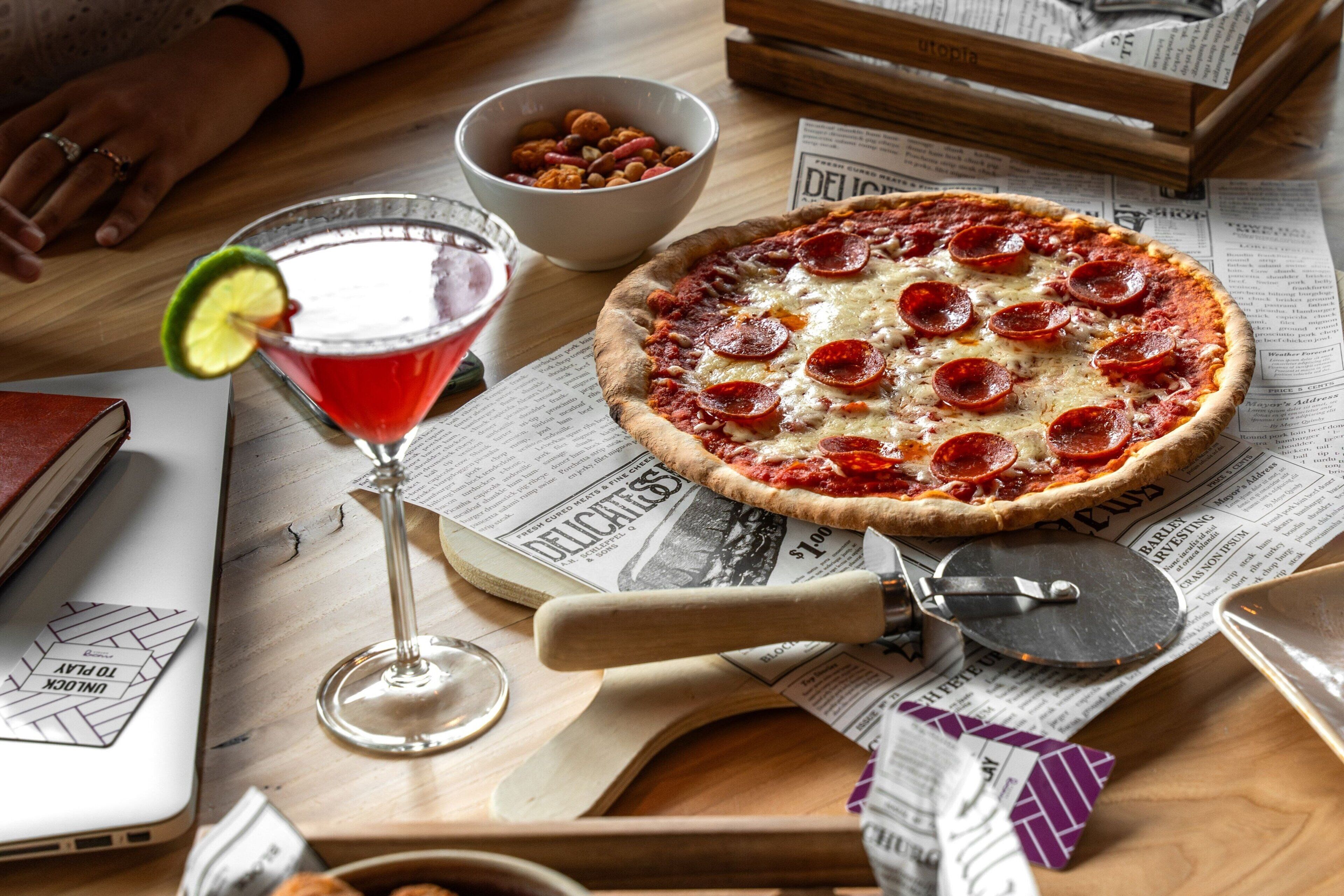 Una pizza sobre un periódico, una copa de martini con lima y un cortador de pizza sobre una mesa de madera.