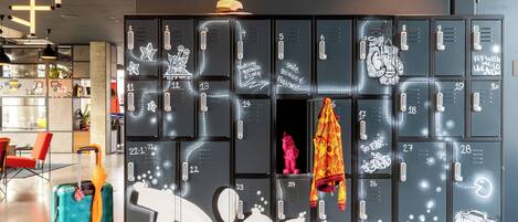 A row of lockers with numbers and motivational quotes, a turquoise suitcase with an orange umbrella, and a red chair in a modern lobby.