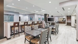 A modern hotel dining area with multiple tables and chairs, a kitchen in the background, and a large flat-screen TV mounted on the wall.