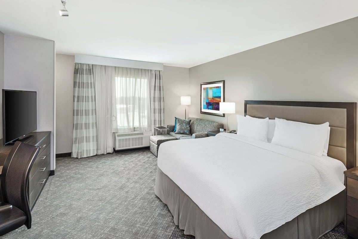 Ein Hotelzimmer mit einem großen Bett, einem Schreibtisch mit Stuhl, einem Fernseher, einer Lampe und einem Fenster mit Vorhängen.