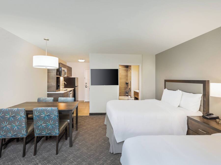 Quarto de hotel com duas camas, uma mesa de jantar com cadeiras, uma kitchenette e uma TV fixada na parede.