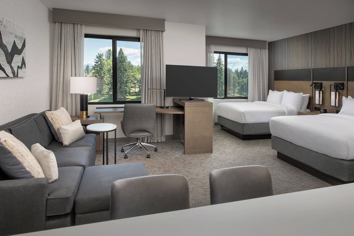 A hotel room with a sofa, two beds, a desk with a TV, and a view of trees.