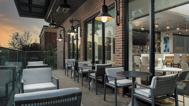 An outdoor seating area with wicker chairs and tables, large windows, and pendant lighting.