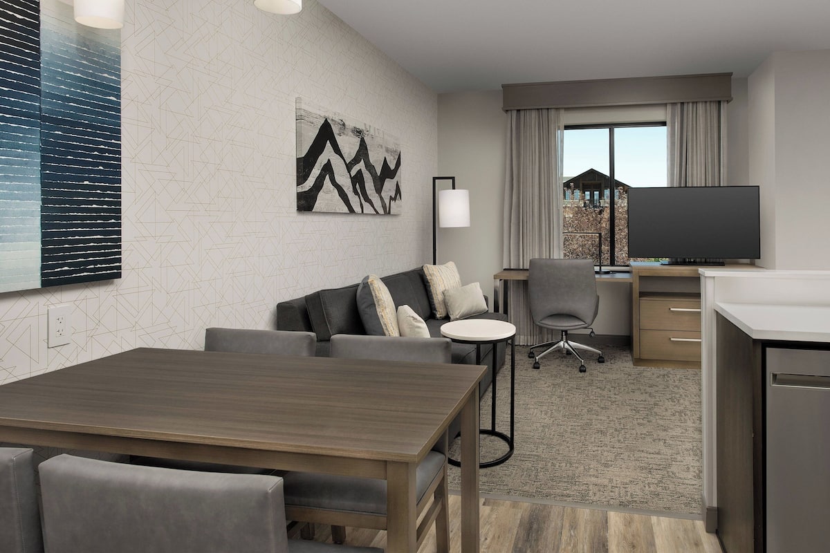 A modern hotel room with a dining area, a sofa set, a flat-screen TV, and a large window with curtains.