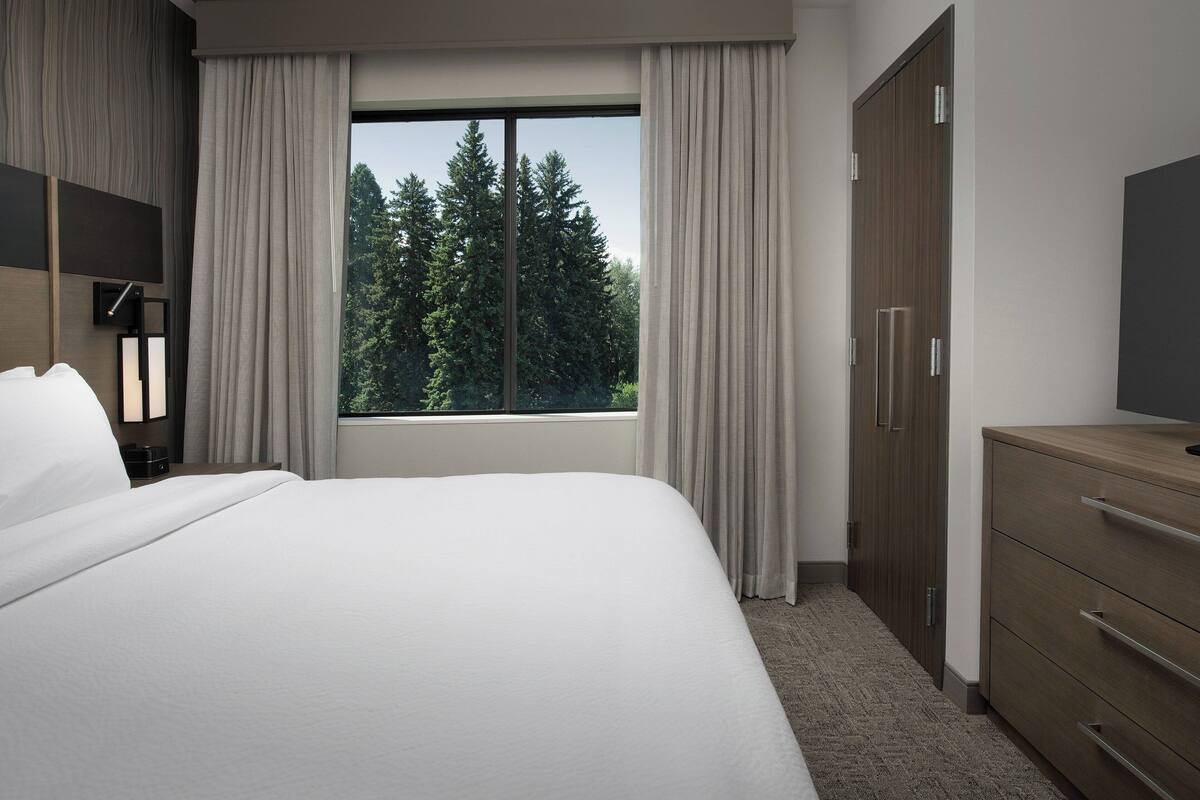 A hotel room with a large bed, a wardrobe, a desk with a lamp, and a window with a view of trees.