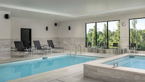 A modern indoor swimming pool with clear blue water, surrounded by lounge chairs and a view of greenery through large windows.