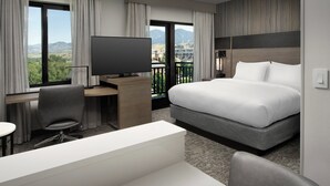 A hotel room with a bed, a desk with a TV, a chair, and a view of mountains through large windows.
