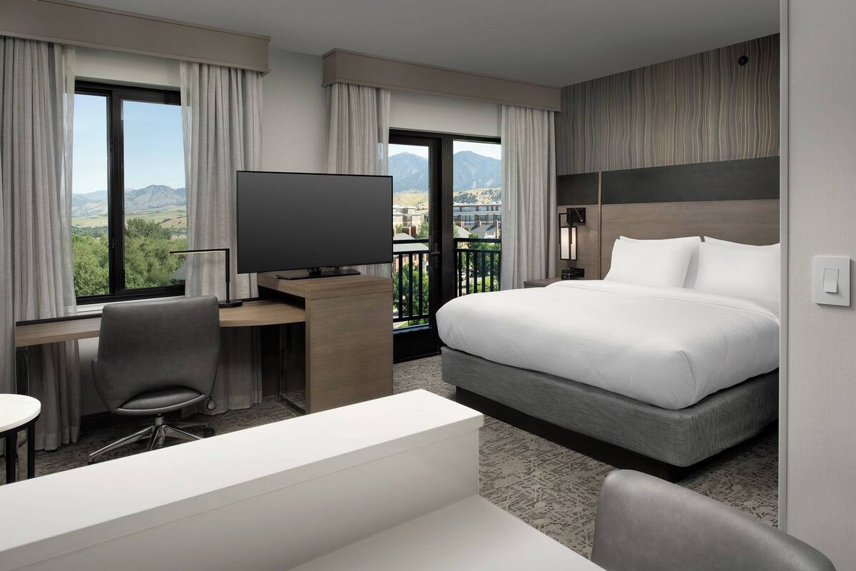 A hotel room with a bed, a desk with a TV, a chair, and a view of mountains through large windows.
