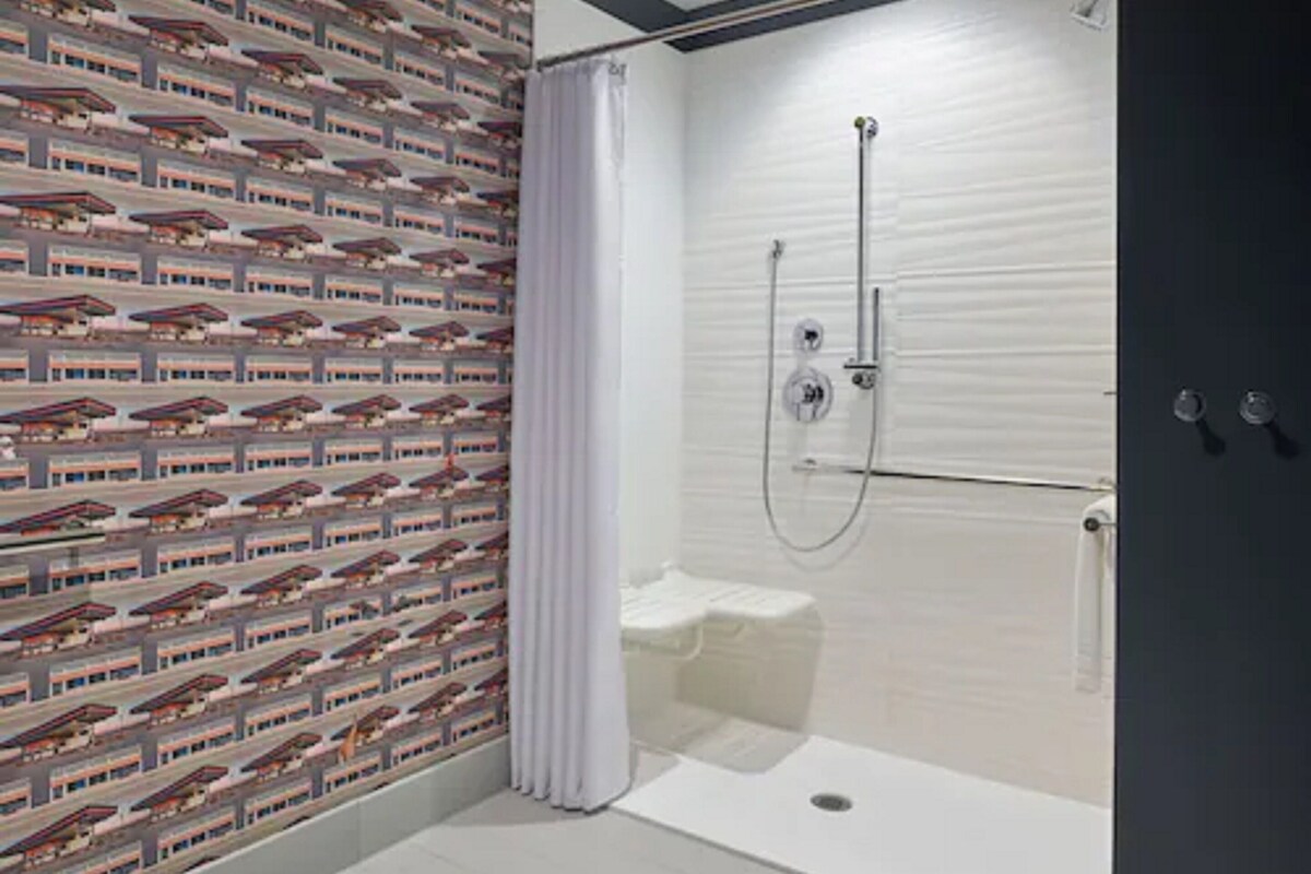 A shower with a glass door, a white curtain, and a tiled wall with a repeating pattern.
