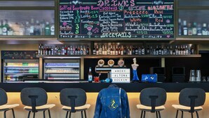 A bar area with a chalkboard menu listing drinks and mocktails, a selection of bottled beverages, and a neon sign. - Moxy Hamburg Altona (Hamburg)