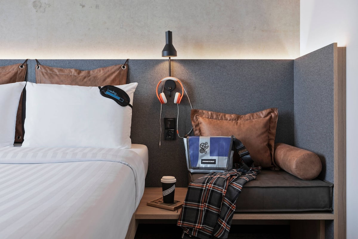A hotel room with a bed, a bedside table with a coffee cup, headphones, and a pillow with a tag.