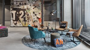 A modern lounge area with comfortable seating, a large abstract wall art piece, and a view through a glass door. - Moxy Hamburg Altona (Hamburg)