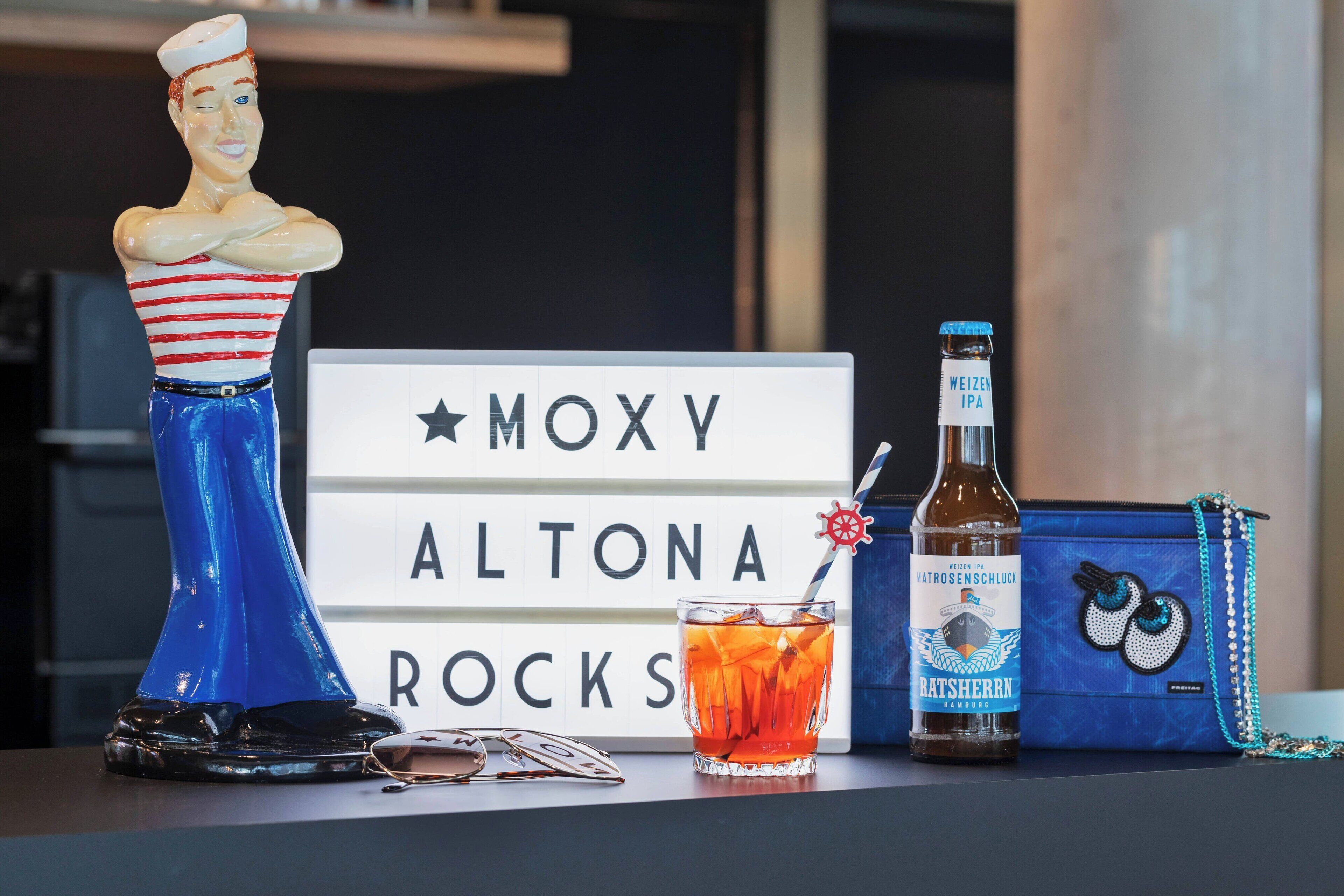 A table with a decorative figure, a sign reading "MOXY ALTONA ROCKS", a bottle of Weizen IPA, a glass of beer, and a blue bag with a chain strap.