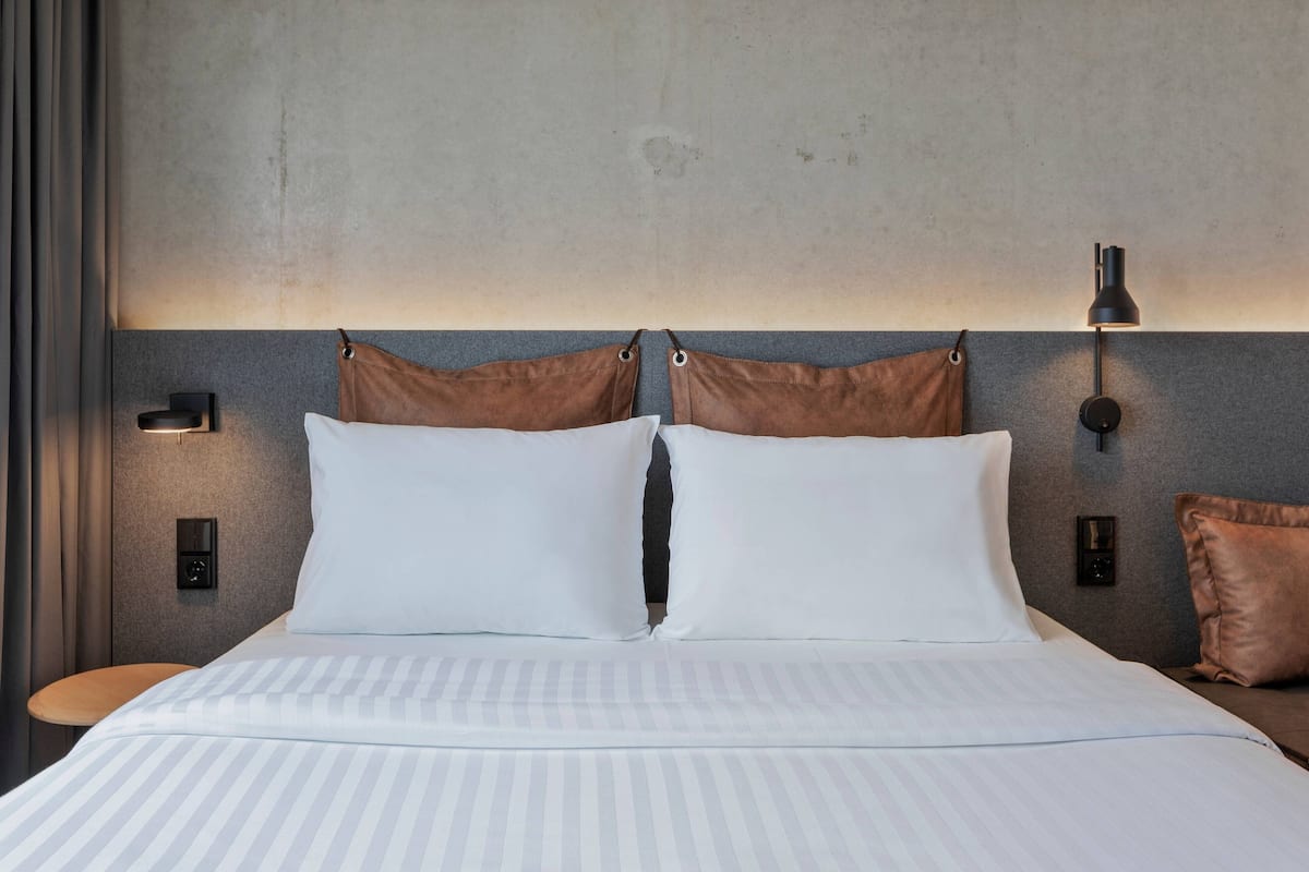 A neatly made bed with white linens and two brown pillows, a bedside table, and a wall-mounted lamp.