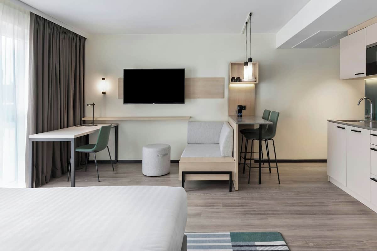 A modern hotel room with a bed, a desk with a chair, a TV mounted on the wall, a small kitchenette, and a window with curtains.