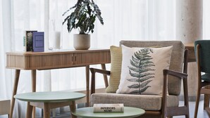 A modern living room with a wooden table, a green round table, a sofa with a fern pillow, a potted plant, and books on a side table. - Residence Inn by Marriott Hamburg Altona (Hamburg)