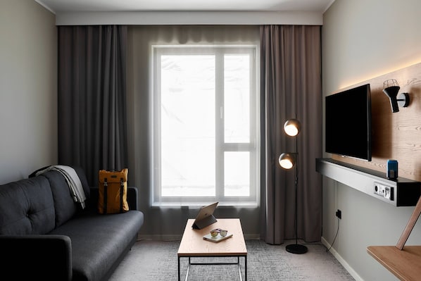 A modern living room with a sofa, a coffee table with a laptop, a TV mounted on the wall, and a floor lamp. - Moxy Milton Keynes (Milton Keynes)